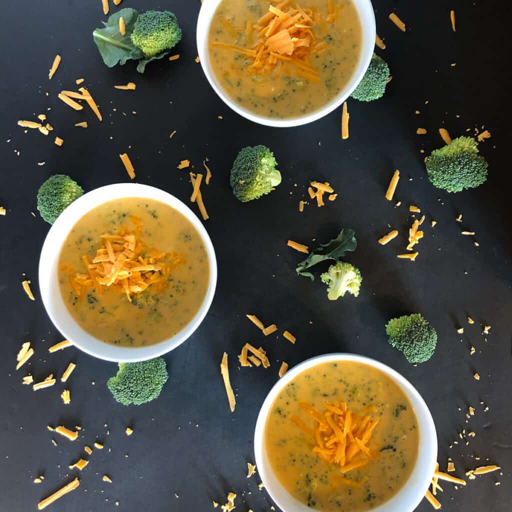 Various Bowls of Broccoli Cheddar Soup Surrounded by broccoli and shredded cheese