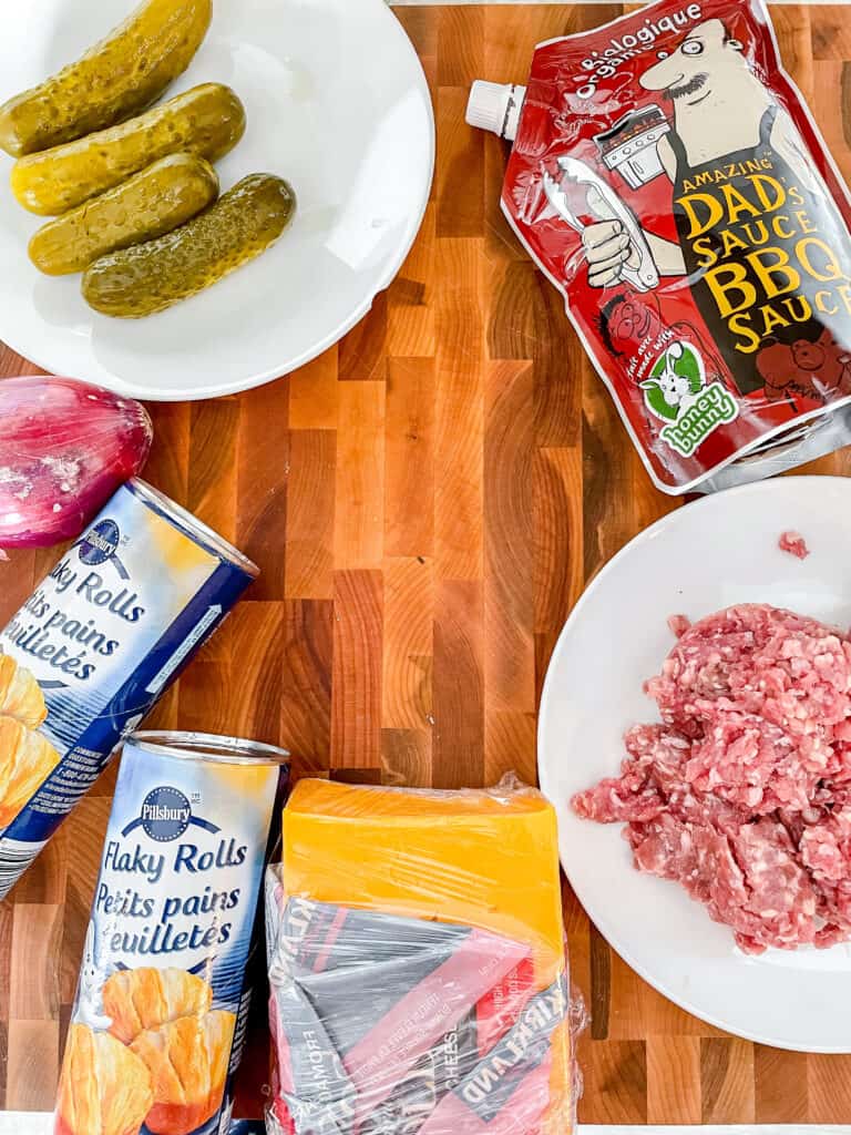 Ingredients for cheeseburger cups on a cutting board. Ground beef, pickles, onion, cheese, sauce and dough