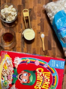 ingredients for lucky charm marshmallow bars. Box of lucky charms, butter, salt, vanilla extract, mini marshmallows and white chocolate candy melts. All sitting on top of a wooden butcher block