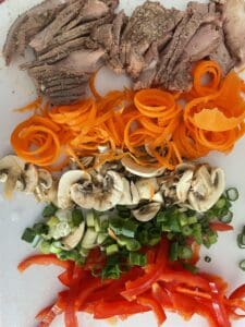 Ingredients chopped up on a cutting board for steak raman, Thinly sliced steak, spiralized carrots, thinly sliced mushrooms, sliced green onion and julienned red bell pepper