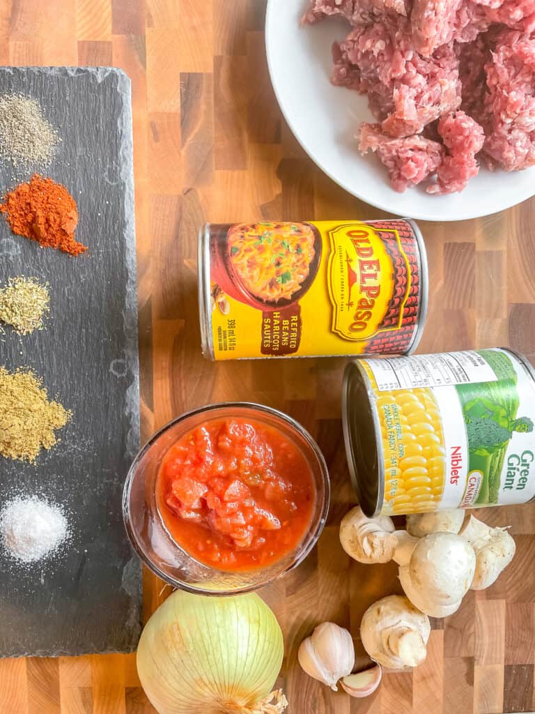 Ingredients to make Beef and Bean Burritos on a wooden cutting board. Raw ground beef on a plate, a can of refried beans, a can of corn, 4 white button mushrooms, garlic cloves, a yellow onion, salsa, and various spices on a piece of black slate.