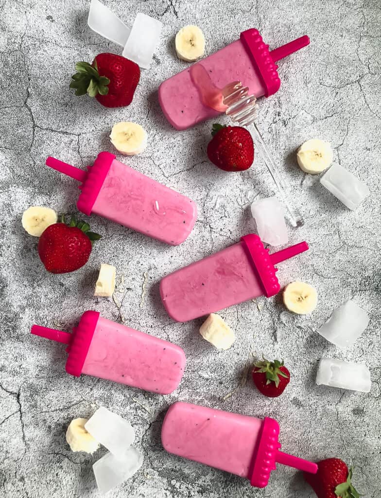 Pink popsicles with pink holders are laying at different angles with strawberries and bananas around them