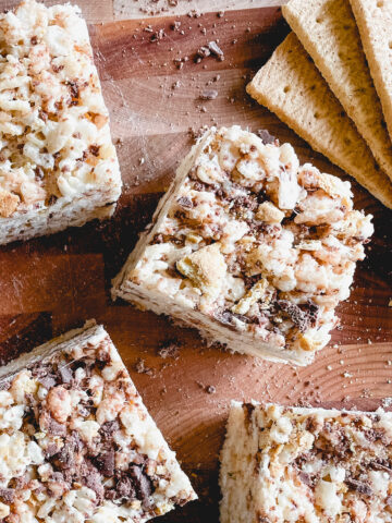 four s'mores rice krispie squares on a wooden cutting board with graham crackers in the background