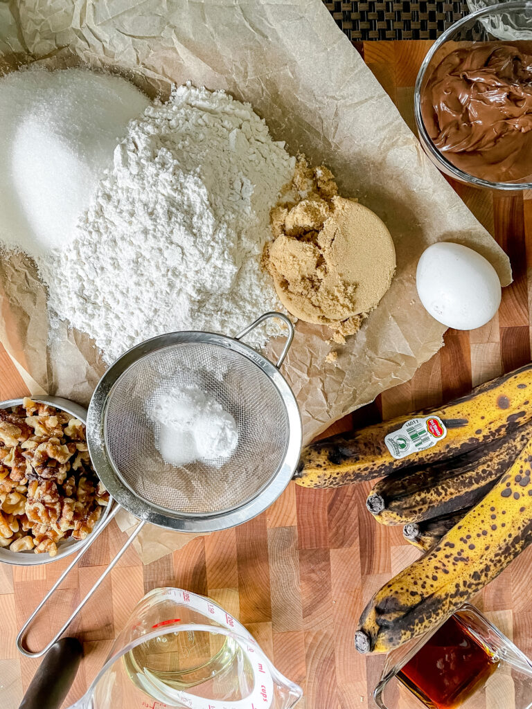 Ingredients for Banana Muffins with Nutella. On a butcher block sits: ripe bananas, oil, flour, sugars, and egg, nutella and leavening agents