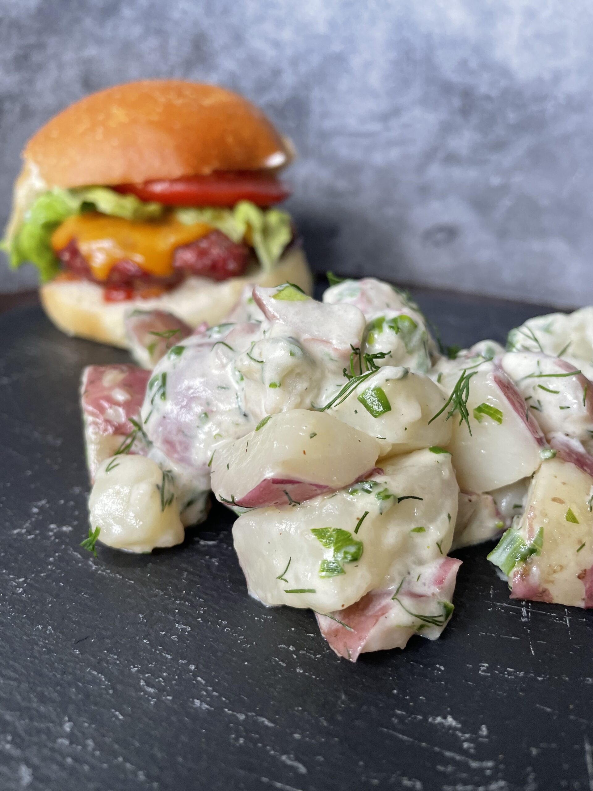 creamy dill potatoes on a black slate plate with a burger in the background